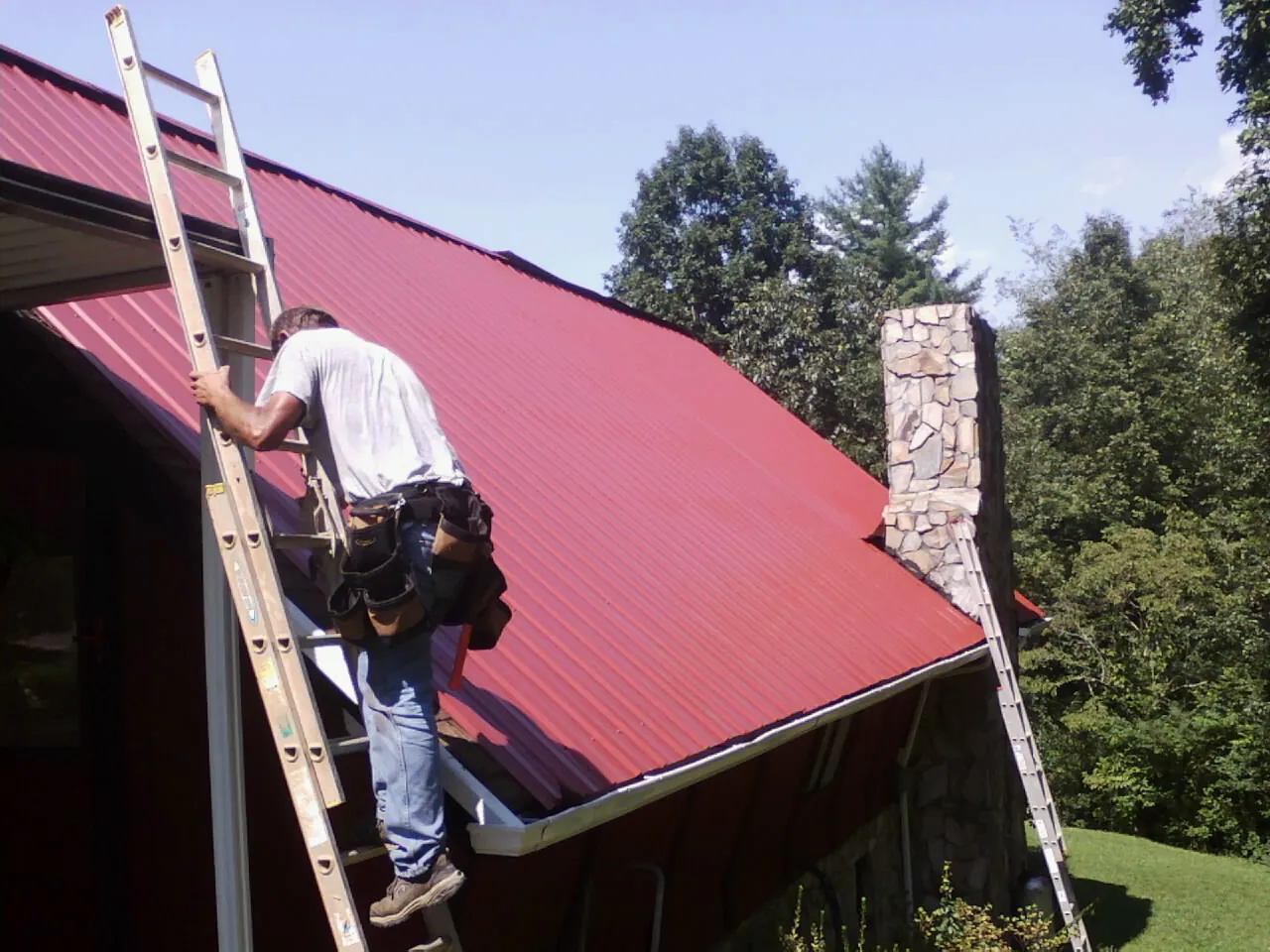 Roofer on a ladder beside a red metal roof and stone chimney — Soffit & Fascia Repair by Roofing Brothers in Dixon